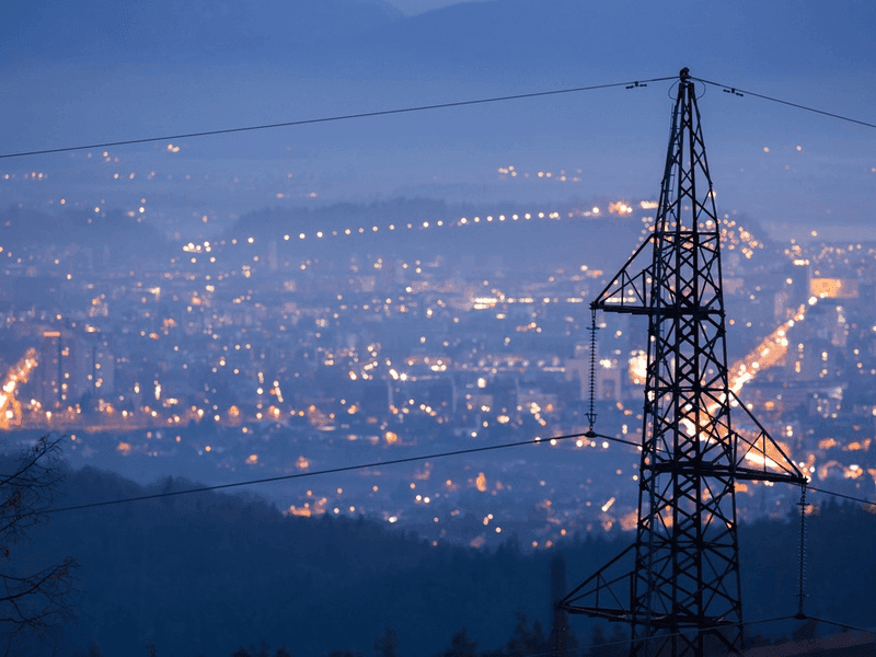 A steel power-line pylon silhouetted at dusk above a valley city glowing with lights.